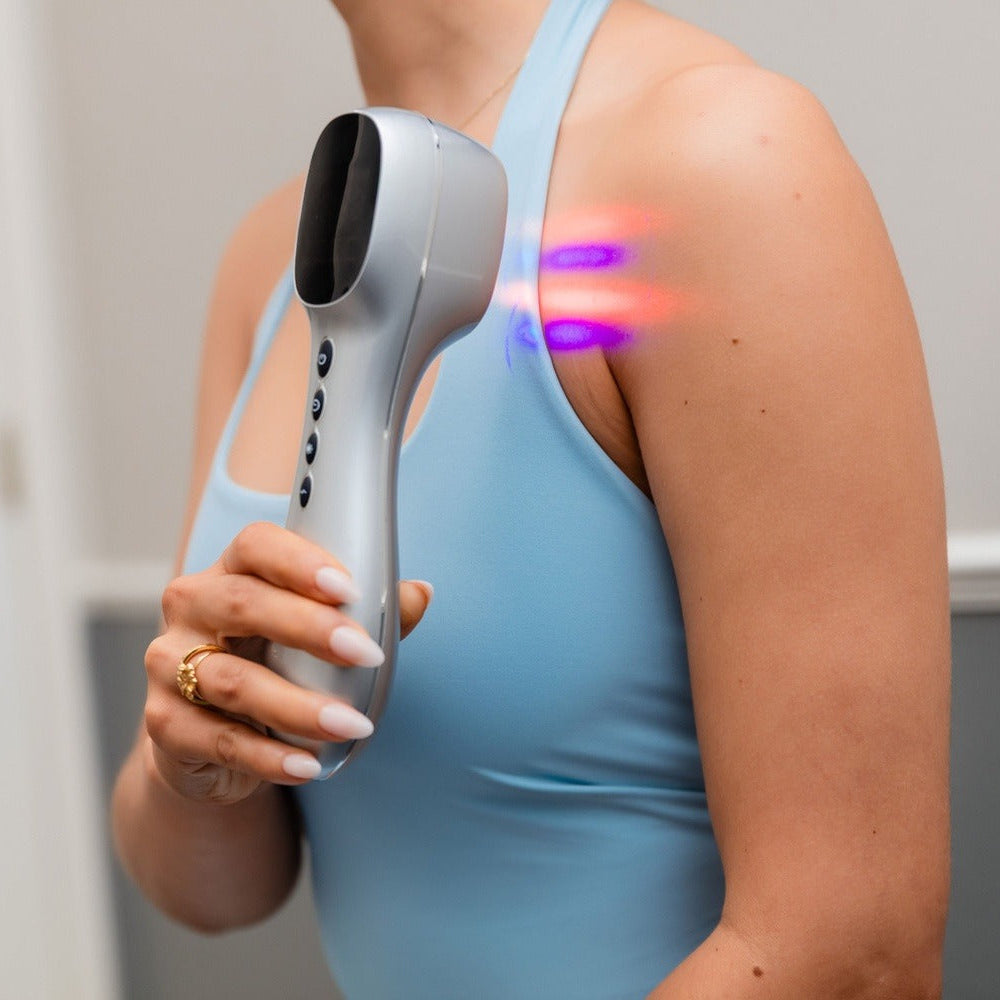 A woman in a light blue tank top uses a handheld silver LED light therapy device on her upper shoulder, with purple and pink light beams visible. The background is a clean, neutral indoor wall.