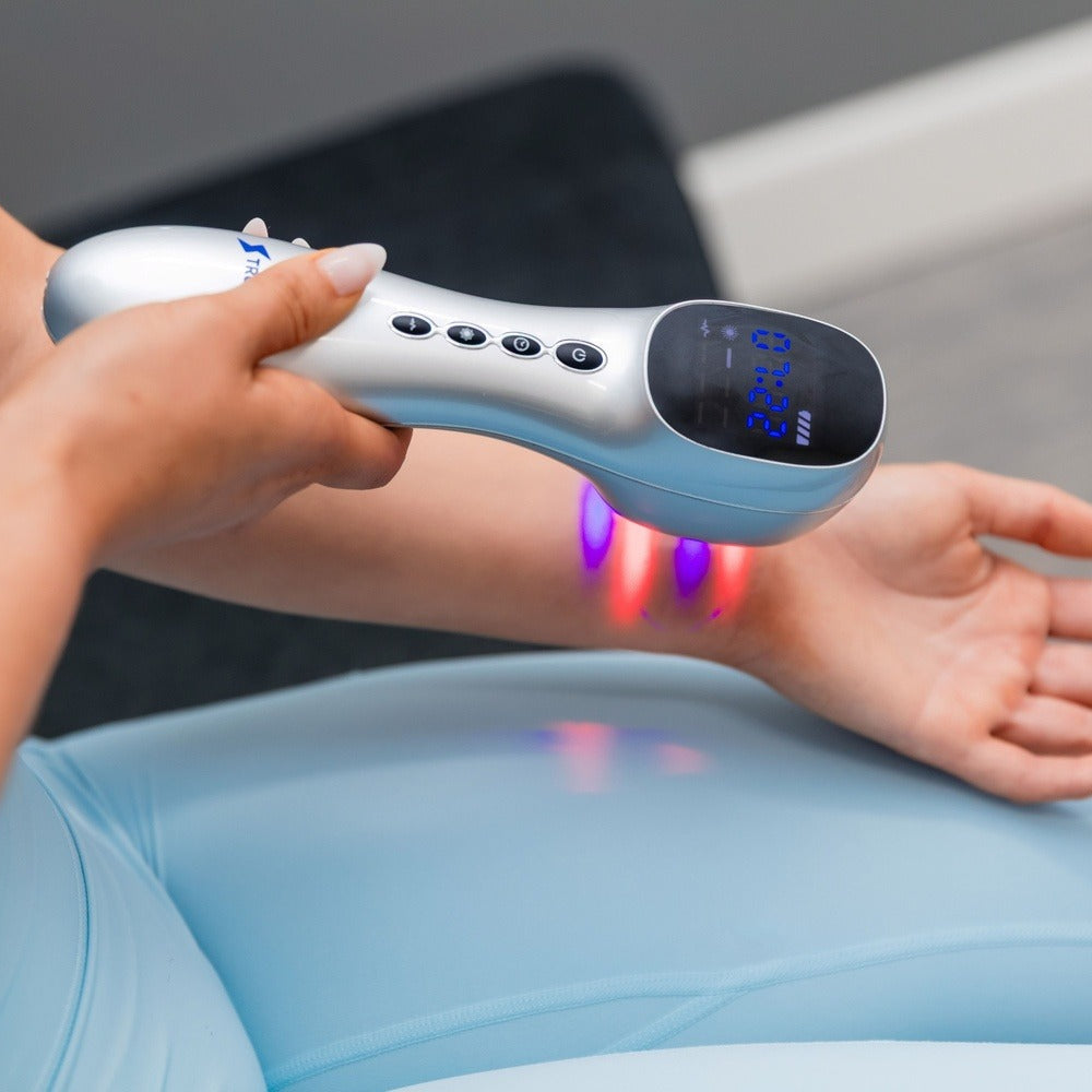 A close-up of a person using a handheld silver and black LED light therapy device on their forearm, with the device emitting red and purple light onto the skin.